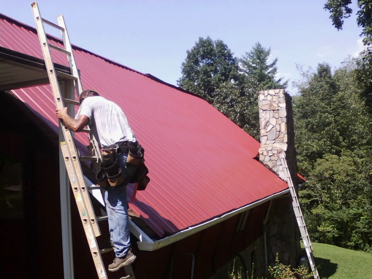 Roofer on a ladder beside a red metal roof and stone chimney — Soffit & Fascia Repair by Roofing Brothers in Mount Ephraim